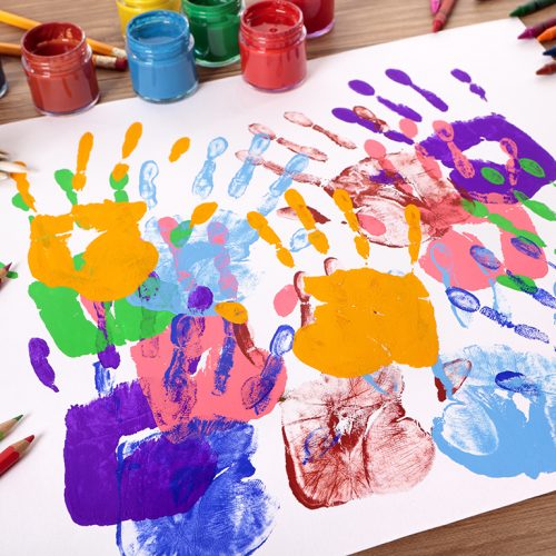 Painted handprints with art and craft equipment on a school table.  Shallow depth of field with sharp focus on the nearest handprints and blurred distant background.