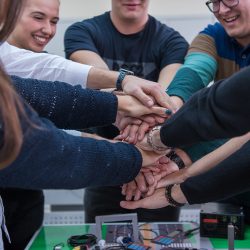 Group of young students in electronics classroom celebrating successfully finished project with holding their hands together, education and technology concept